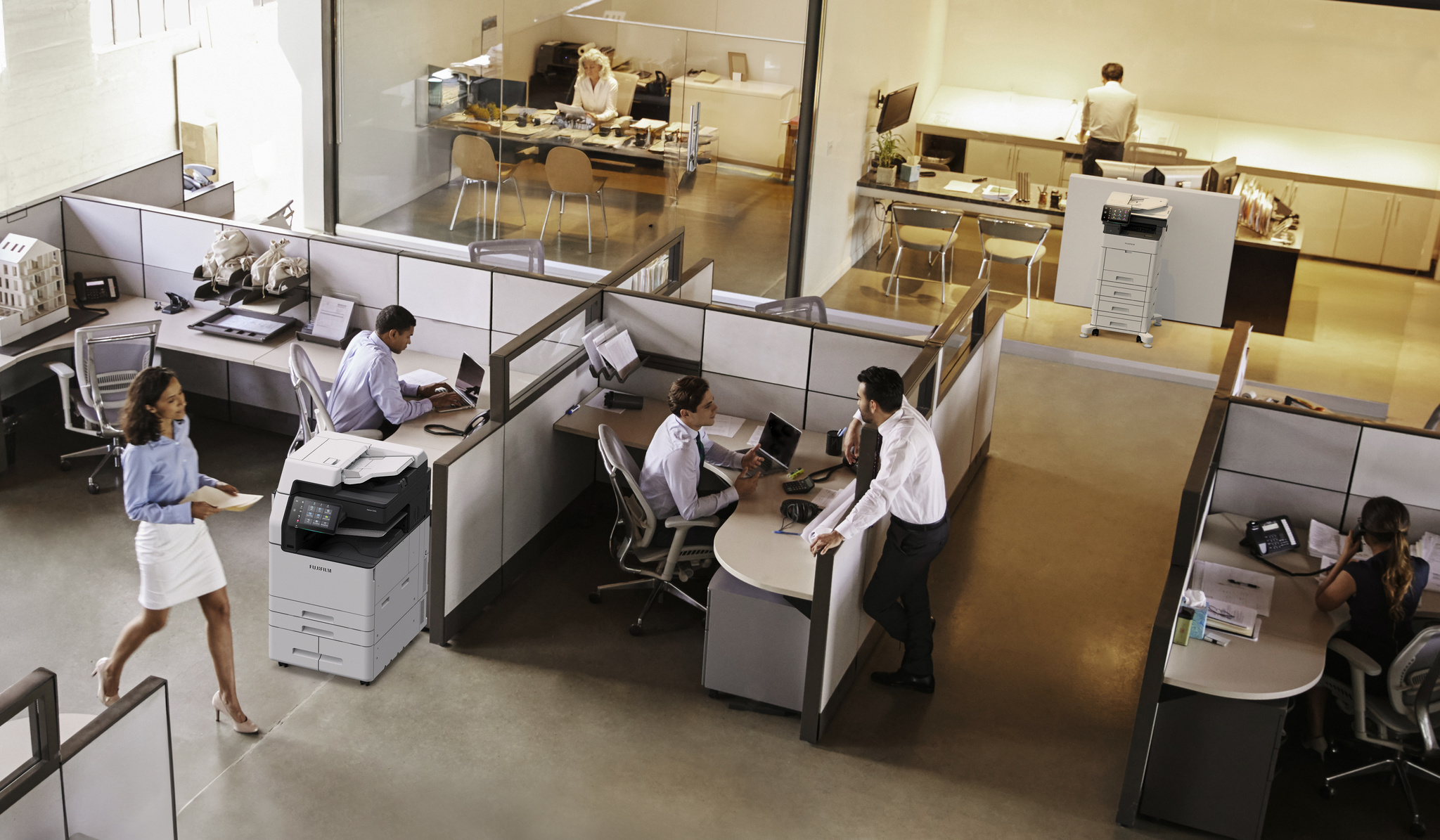 Open-plan office with employees working in cubicles and meeting areas, with FUJIFILM multifunction printers placed near workstations.